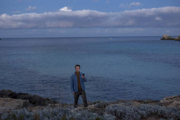 Man holding smart phone standing on hill near sea at dusk - JOSEF23656