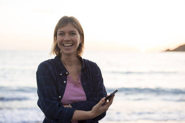 Fröhliche Frau mit Smartphone am Strand bei Sonnenuntergang - JOSEF23675