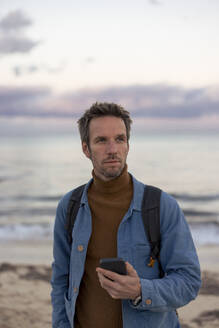 Thoughtful man holding smart phone at beach - JOSEF23689