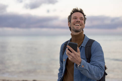 Carefree man holding smart phone at sunset beach - JOSEF23692