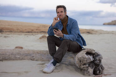 Thoughtful man holding smart phone sitting on driftwood at sandy beach - JOSEF23694