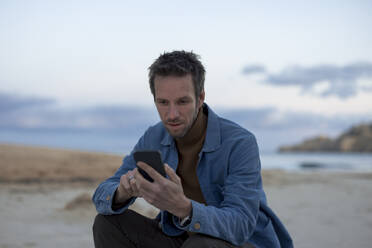 Mann benutzt Smartphone bei Sonnenuntergang am Strand - JOSEF23695