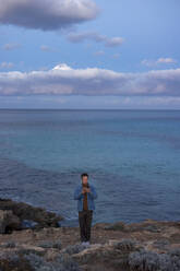 Man using smart phone standing on hill near sea at dusk - JOSEF23698