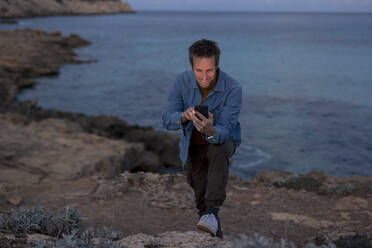 Smiling man using smart phone on hill near sea at dusk - JOSEF23700