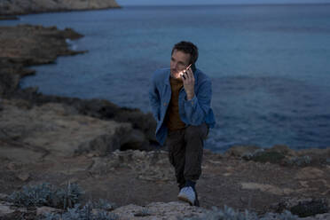 Man talking on smart phone on hill near sea at dusk - JOSEF23701