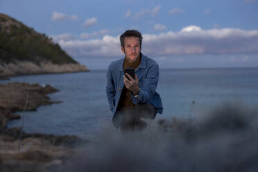 Man with smart phone on hill near sea at dusk - JOSEF23702