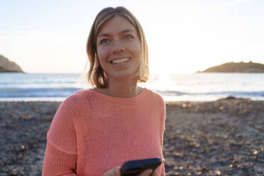 Glückliche Frau mit Handy in der Hand verbringt ihre Freizeit am Strand bei Sonnenuntergang - JOSEF23718