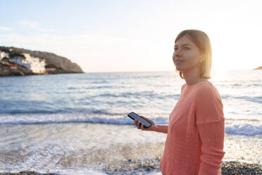 Thoughtful woman with smart phone on shore at beach - JOSEF23719