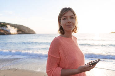 Lächelnde Frau hält Smartphone am Ufer bei Sonnenuntergang Strand - JOSEF23720