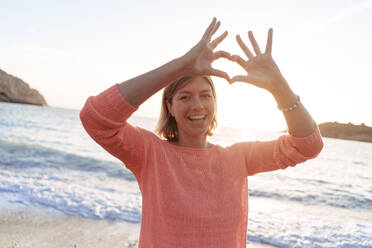 Happy woman gesturing heart shape at sunset beach - JOSEF23722