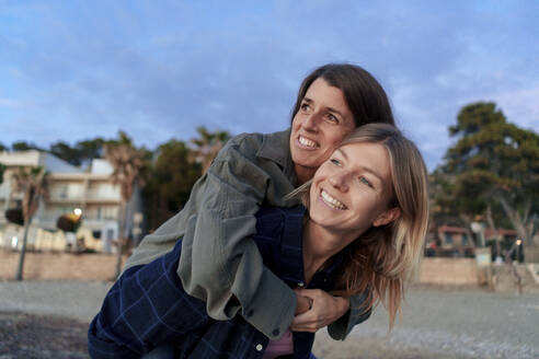 Happy woman piggybacking friend at sunset beach - JOSEF23733