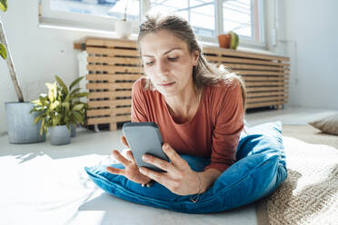 Lächelnde Frau, die auf einem Kissen liegt und ihr Smartphone zu Hause benutzt - JOSEF23771