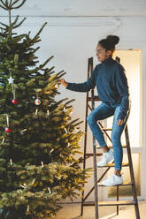 Young woman decorating Christmas tree standing on ladder - JOSEF23778