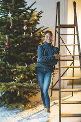 Woman standing by ladder and Christmas tree at home - JOSEF23780