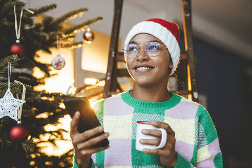 Nachdenkliche Frau mit Smartphone und Kaffeetasse zu Hause - JOSEF23812