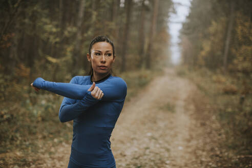 Pretty young woman in blue track suit stretching before workout in the autumn forest - INGF12990