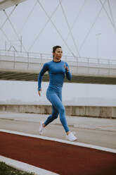 Pretty young woman in blue track suit running by the river at autumn morning - INGF12997