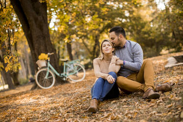 Cheerfull young couple sitting on ground in autumn park - INGF12999