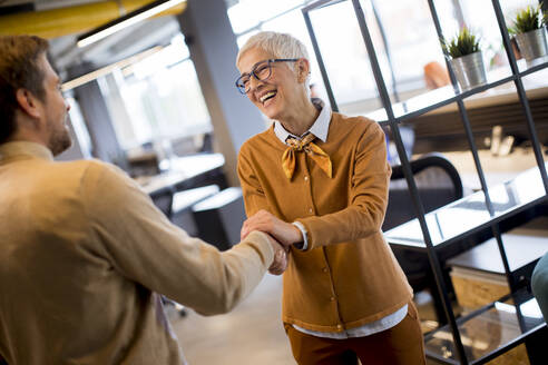 Grateful senior businesswoman boss lady hold hand of best employee, shake hands warm welcoming standing in coworking space - INGF13046