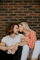 Handsome ymiling young couple in love sitting in front of house brick wall - INGF13084