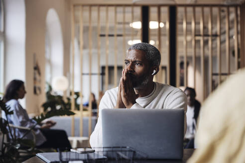 Worried businessman with hands clasped talking through wireless in-ear headphones at office - MASF43004