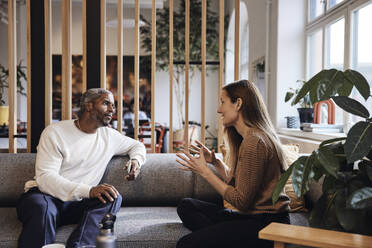 Businesswoman discussing with male colleague while sitting on sofa at coworking office - MASF43021