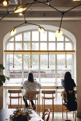 Rear view of businesswomen working on laptop while sitting on chair near window at coworking office - MASF43040