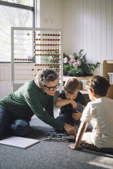 Senior female teacher explaining preschool boys sitting in classroom at preschool - MASF43050