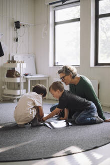 Schoolboys studying with senior female teacher while sitting on classroom floor at preschool - MASF43052