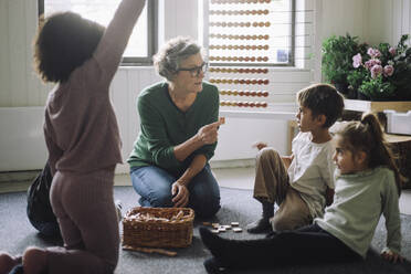 Senior female teacher playing puzzle game with preschool children while sitting in classroom at kindergarten - MASF43058