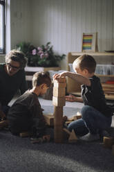 Schoolboys playing with toy blocks with female teacher in classroom at kindergarten - MASF43065