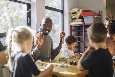 Smiling male teacher holding clay while sitting with kids in classroom at preschool - MASF43070