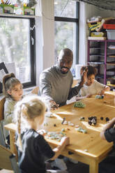 Male teacher playing with kids while sitting at bench during art class at preschool - MASF43071