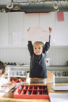 Portrait of girl showing paper with drawing while standing near bench in classroom - MASF43076