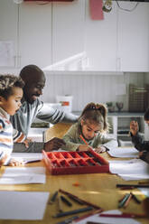 Smiling male teacher assisting students writing on paper while sitting in class at kindergarten - MASF43079