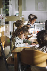 Girl writing on paper while sitting with students at bench in classroom - MASF43081