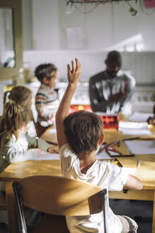 Rear view of boy raising hand while sitting on chair in classroom at kindergarten - MASF43082