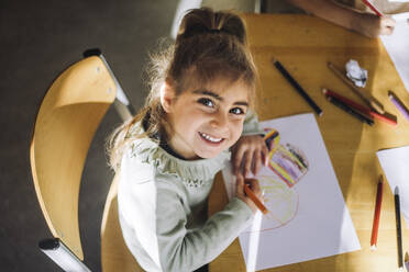 High angle view of smiling girl drawing with crayon on paper while sitting at bench in classroom - MASF43083