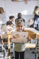 Portrait of smiling girl showing drawing while standing near bench in classroom at kindergarten - MASF43084