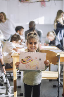 Portrait of smiling girl showing drawing while standing near bench in classroom at kindergarten - MASF43084