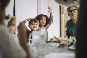 Children raising hands while sitting with female teacher during lunch time at kindergarten - MASF43087