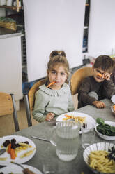 Portrait of playful girl holding carrot slice in mouth while sitting at dining table during lunch time - MASF43091