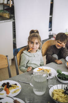 Portrait of playful girl holding carrot slice in mouth while sitting at dining table during lunch time - MASF43091