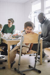 Portrait of smiling boy sitting on chair with teacher at dining table during lunch break - MASF43094