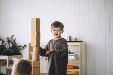 Boy looking away while standing near tower of toy blocks in classroom at kindergarten - MASF43102