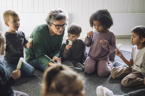 Group of children eating popsicle with senior female teacher while sitting in classroom at kindergarten - MASF43104