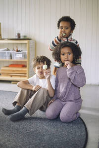 Portrait of boys and girl eating ice cream while sitting on carpet in classroom at kindergarten - MASF43105
