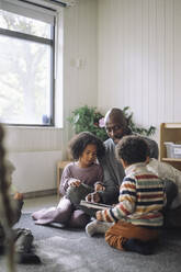 Male teacher reading to preschool kids while sitting in classroom at kindergarten - MASF43106