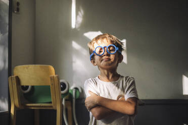 Portrait of boy wearing blue eyeglasses and standing with arms crossed in classroom at kindergarten - MASF43118