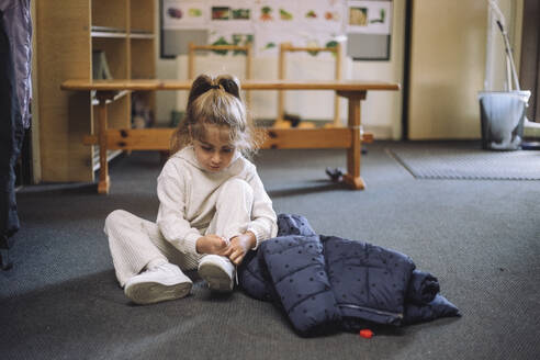 Girl tying shoelace while sitting with jacket on floor in classroom at kindergarten - MASF43121
